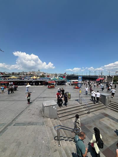 The image depicts a bustling pedestrian area near the Galata Bridge in Fatih, Istanbul, Turkey, under a clear blue sky with scattered white clouds. The foreground features a wide, paved plaza with intricate stone tile patterns, leading to a set of concrete stairs. Several individuals are visible walking up and down the stairs, some in casual attire, while others are dressed in headscarves.
To the left, a crowd congregates around food stalls set up along a waterfront promenade. These stalls, adorned with red awnings, appear to be selling local cuisine. People are seen ordering, eating, and socializing around tables. Further back, a body of water is visible, with boats and ferry terminals lining the shore. Beyond the water, the iconic skyline of Istanbul, including the Galata Tower, is visible.
In the background, the Galata Bridge spans the water, with vehicles and pedestrians crossing it. A tram line runs along the bridge, indicated by signage. The overall scene conveys a sense of vibrant urban activity and tourism, characteristic of a popular landmark in Istanbul. The sunlight casts distinct shadows on the ground, indicating it is midday.