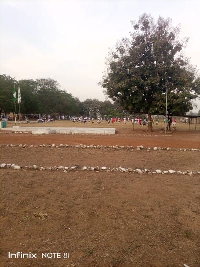 A wide shot captures a field with a sparse, brown surface, bordered by rows of white stones. In the background, a soccer game unfolds with players in white and red uniforms. A large tree dominates the right, partially obscuring the view of the game. To the left, a flag stand displays two flags. The sky is overcast, suggesting a dull day. "Infinix NOTE 8i" is visible at the bottom.