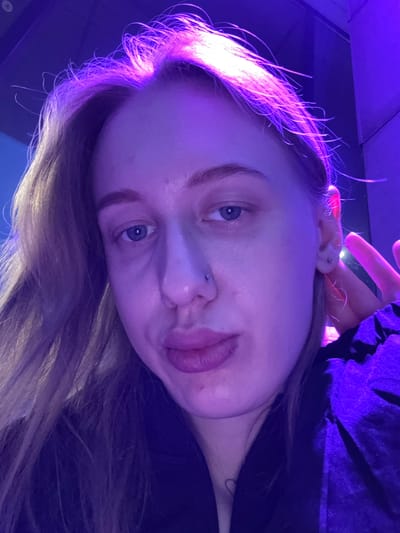A close-up, angled photograph features a light-skinned person with blonde/brown hair, possibly female, under purple and blue lighting. The person has blue eyes and a nose piercing. Their tongue is slightly protruding from their mouth. Part of their hand is visible near their ear, with a possible piercing. The background is dark and indistinct, suggesting an indoor setting. No specific details indicate the location of Ol'ginskaya, Russia.