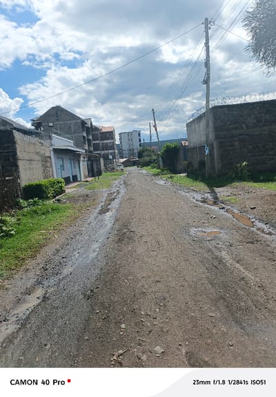 The image depicts a unpaved dirt and gravel road in Nakuru East ward, Kenya, showing evidence of recent moisture with several muddy puddles and wet patches. The road, which shows tire tracks, extends from the foreground into the middle distance, curving slightly right.
On the left side of the road, an area of green grass borders a low, light-colored building with light blue trim around its windows or doors, partially obscured by bushes. Behind this structure, a concrete wall topped with coiled razor wire extends. Further along this side, multiple multi-story residential or commercial buildings are visible, some appearing to be under construction with exposed concrete blockwork, while others have reddish-brown roofs. A white vehicle is faintly visible parked further down the road on the left.
On the right side, a substantial stone wall, also topped with coiled razor wire, runs parallel to the road. Beyond this wall, an electric utility pole with multiple cross-arms, insulators, power lines, and associated equipment stands prominent. Another similar utility pole is visible further down the road. Patches of green vegetation are scattered along both sides of the road and in the background.
The sky overhead is partly cloudy, with significant areas of blue sky interspersed with grey and white clouds, suggesting a daytime scene. No individuals, direct actions, or interactions are visible within the frame. The overall environment suggests an urban or semi-urban area undergoing development.