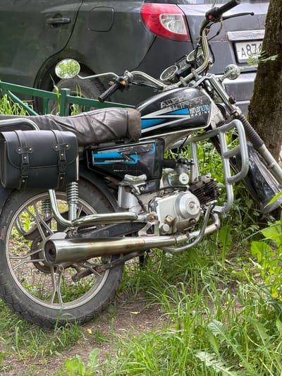 An outdoor shot depicts a parked motorcycle next to a dark-colored car, set against a backdrop of green grass, weeds, and a tree. The photograph appears to be taken in daytime, with natural light illuminating the scene.

The motorcycle, positioned on the right side of the frame, is a black model with blue and white graphics on its fuel tank. Visible text on the tank reads "HIFILFILTRO RACING" and "ALEX". It features silver handlebars with two round gauges, a small circular mirror (reflecting green foliage), and a silver engine block with an exhaust pipe. A black, leather-like saddlebag with buckles is attached to the side, and a matching material, possibly part of the seat or another bag, is draped nearby. The front wheel, with a silver rim and black tire (faintly showing "TUBELESS" and "INDIA" on the sidewall), is visible towards the bottom right. Chrome-plated crash bars frame the front of the motorcycle.

On the left, partially obscured by the motorcycle and a tree trunk, is the rear section of a dark car, likely an SUV or van. Its red taillight is visible, along with a rear license plate displaying "A874". A portion of the car's tire and a black handle are also discernible.

The ground is covered with dense green grass and various weeds, with some bare soil showing through. A large tree trunk is visible in the upper part of the image, contributing to the natural, slightly overgrown setting. A green metal structure, possibly a fence or gate, can be seen in the lower left, behind the car. The location is Tokkari, Russia, as suggested by the car's license plate style.