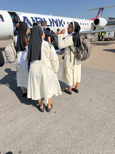 In Kempton Park, South Africa, three nuns in traditional white habits and black veils are standing on an airport tarmac. One nun, facing left, has a colorful scarf around her neck and a light-colored backpack slung over her shoulder. The other two nuns are facing away from the camera, with one of them in the center and another on the right. Both have backpacks. Behind them, a white Airlink airplane with blue lettering is parked, and passengers are boarding or disembarking through the open door. The plane's tail features a colorful logo. In the background, airport personnel in high-visibility vests are visible, along with airport equipment and structures. The scene is illuminated by bright sunlight, casting shadows on the concrete ground.
