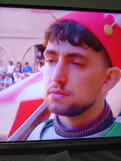 The image shows a close-up of a man's face wearing a red cap with a gold emblem and a white feather. He has dark hair and a beard, and is wearing a costume that appears to be part of a historical reenactment or parade. In the background, there are blurred figures of people seated in what appears to be an outdoor setting, possibly a piazza in Siena, Italy, given the context of historical reenactments often associated with the city. The man is holding a red and white striped banner. The overall impression is that of a participant in a local festival or historical event.
