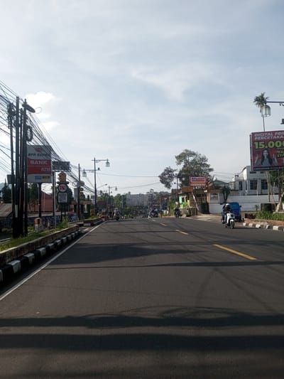 The image depicts a street in a town with a clear sky above. The road is paved and has a yellow dividing line. There are sidewalks on either side.
Various signs are visible on the roadside, including one for "BPR Supra Bank ATM" and another for "Alfamart". There is a billboard on the right side of the road. Overhead, there are numerous power lines and street lights. Several motorcycles are traveling on the road. The location appears to be a town or small city in Indonesia or southeast Asia, characterized by narrow streets, tropical vegetation, and small businesses along the roadside. It is not located in Phou Khoun, Laos.