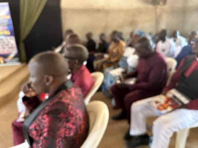 The image depicts a gathering of people, likely in a church or community hall in Kufana, Nigeria. The setting appears to be indoors, with a plain, light-colored wall and a dark curtain or drape visible in the background. The lighting suggests it is daytime.

The subjects are predominantly adults seated on chairs, facing forward as if attending a service or presentation. The image is out of focus, making it difficult to discern individual features with clarity. However, several individuals are wearing brightly colored attire, including a man in the foreground with a dark jacket featuring a vibrant red floral pattern, and another person in a maroon garment. Others are dressed in lighter colors, such as white shirts and light blue head coverings.

One person in the foreground is holding a booklet or magazine. The overall atmosphere appears to be one of attentive attendance. There is no clear indication of the specific activity or event beyond the fact that people are gathered and listening. There is some blurry text visible on a banner-like object in the far left of the image, but it is too indistinct to read.