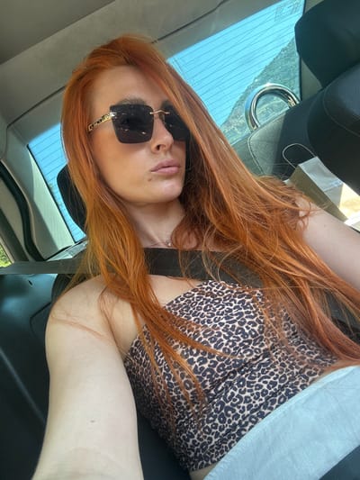 A woman with long, reddish-brown hair is captured in a selfie from an elevated perspective, lying down in what appears to be the passenger seat of a car. She is wearing a leopard-print top and large, square-shaped, dark-lensed sunglasses. Her head is tilted, and her gaze is directed slightly away from the camera.
The background includes a car's interior, with the headrest of the seat behind her visible. Through the car window, a bright sky and lush green hills are discernible, suggesting a daytime setting with clear weather. The overall impression is one of a relaxed moment, possibly during a road trip.
The visible text, partially obscured on a white paper bag in the background, reads "MANDIĆ PHARMACY". The location context of Mišići, Montenegro, aligns with the scenic greenery seen through the window.