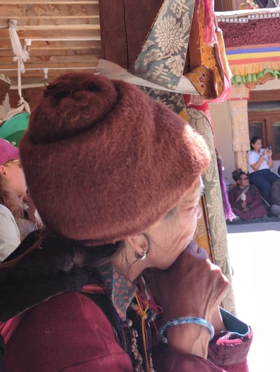 The image shows an outdoor scene, possibly in Pibiting, India, with a focus on an elderly woman in the foreground. She is wearing a large, fuzzy, reddish-brown hat and a maroon jacket with a patterned collar. Her right hand is raised to her face, with a light blue, patterned bracelet on her wrist. To her left, partially visible, is another person wearing a pink hat and a white top. In the background, a woman in a white shirt is holding a phone, and two men are seated on the ground. One man is wearing a maroon robe and a traditional hat, and the other is in similar attire. The environment appears to be a courtyard or outdoor gathering area, with decorative fabrics and architectural elements visible.