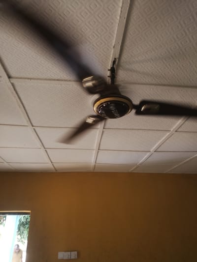 The image shows a ceiling fan in motion, its blades blurred due to the rotation. The fan is mounted on a white, textured ceiling composed of square tiles. The fan itself has a dark body and a metallic accent around the center, where some text is visible. The text on the fan appears to read "AMASE-II".

Below the ceiling, a section of a wall painted in a warm, mustard yellow is visible. To the left of the wall, there is an open doorway or window, revealing a glimpse of greenery and light. A person, possibly a man wearing a brown garment, is partially visible through this opening, suggesting an indoor setting. There are also two light switches or electrical outlets mounted on the yellow wall.

The overall lighting suggests it is daytime. The scene appears to be an interior space, possibly a room in a building. The presence of a ceiling fan and the simple decor hint at a functional, everyday environment. The blurred motion of the fan indicates it is actively running, likely to cool the space. The location context of Ajasse Ipo, Nigeria, suggests a tropical or warm climate where such fans are commonly used.