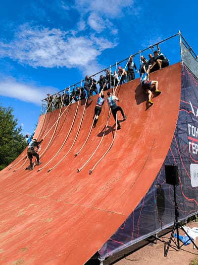 The image depicts an outdoor obstacle course in Sertolovo, Russia. A group of participants are scaling a large, red, curved wall using ropes. Some individuals are near the top of the wall, being assisted by others standing on a metal platform. A few participants are lower down, actively climbing. The environment includes a bright blue sky with scattered clouds, trees in the background, and a dirt ground surface. There is also a speaker on a tripod and some banners with text, one of which reads "Гонка Героев," indicating the event's name, which translates to "Race of Heroes."