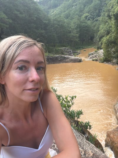 In a selfie taken outdoors, a blonde woman with blue eyes and fair skin is positioned in the foreground, looking directly at the camera. She is wearing a white camisole that exposes her shoulders and décolletage. Her hair is styled in loose waves. Behind her, a murky, brownish-orange river flows through a rocky gorge. The riverbanks are lined with lush green trees and vegetation, with a dense forest extending up the hillsides in the background. The overall setting appears to be a natural landscape, likely in the vicinity of Păng Tiêng 1, Vietnam, given the tropical appearance of the foliage and terrain.