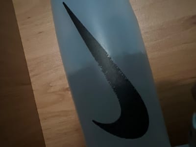 A close-up, overhead shot shows a portion of a light blue Nike water bottle with the black Nike logo on it. The bottle is resting on a wooden surface. The wooden surface appears to be made of planks laid side-by-side, with visible grain and some darker marks. The bottle occupies the left side of the frame, with the logo prominently displayed. The background is out of focus, showing the wooden texture. There are no people or other objects immediately visible in the frame. The scene is well-lit, with the light reflecting off the bottle's surface. The location in the city of Debelt, Bulgaria, is not discernible from the image itself.