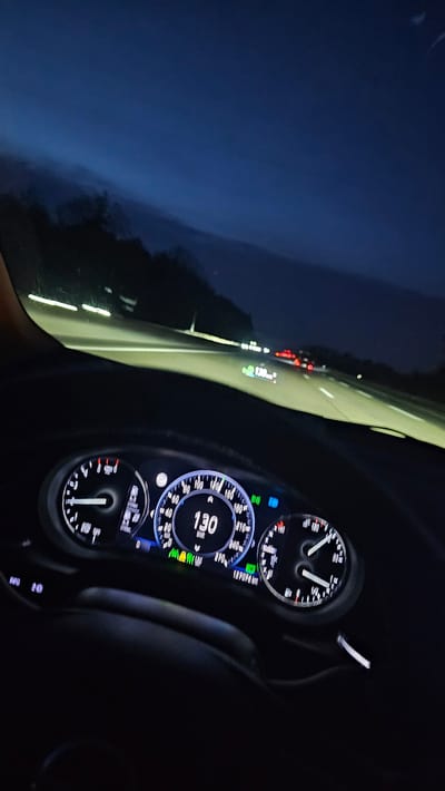 The image depicts the interior perspective of a vehicle, specifically the dashboard and the view through the windshield, at dusk or night, on a multi-lane highway in Seligenstadt, Germany. The vehicle's instrument cluster is illuminated, displaying a speed of 130 km/h on the central digital speedometer. The odometer reads 139,796 km. The tachometer on the left is at a low RPM, and the fuel gauge on the right indicates more than half a tank. Several indicator lights are active, including green icons (likely related to driver assistance systems) and a blue high-beam indicator. The road ahead is illuminated by the vehicle's headlights, revealing a light-colored asphalt surface. In the distance, multiple vehicles are visible, distinguished by their red taillights. Two distinct head-up display projections or digital signs are visible on the road ahead, each displaying "130 km/h," indicating the speed of preceding vehicles or a consistent speed suggestion. Dark silhouettes of trees or vegetation line the highway's edge against a deep twilight blue sky with subtle cloud formations. No individuals are directly visible, but the perspective is from the driver's seat.