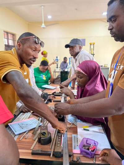 Afaka, Nigeria. A classroom setting. Individuals are gathered around a wooden table. A woman wearing a maroon hijab is observing or participating in a hands-on activity involving metal rods and a mechanical device. The group appears to be engaged in a practical learning exercise. Other individuals are visible in the background, including someone in a green shirt. The table has tools, notebooks, and writing instruments. The scene suggests an educational or training environment.