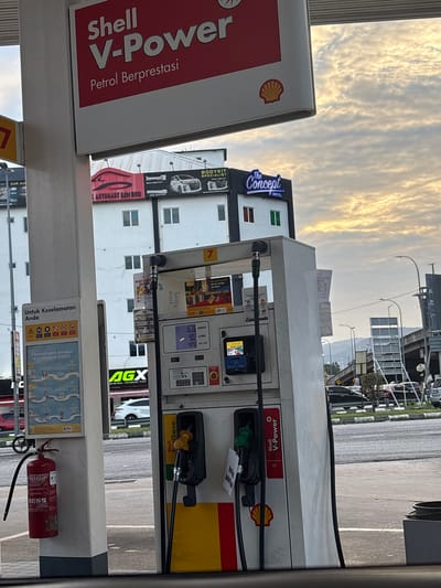 The image shows a gas station in Selayang Municipal Council, Malaysia. The gas station is a Shell station, selling Shell V-Power petrol. A price display shows 11.36 Ringgit and 4.07 litres. A red fire extinguisher is located at the base of the gas pump. In the background, buildings with businesses such as The Concept Hotel and Automart SDN BHD are visible. There are advertising banners for AGX System Window Tint and body kit specialists. Above the building is a cloudy sky.