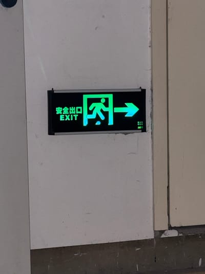 This media file shows a close-up of a vertical exit sign mounted on a white wall. The sign is illuminated in bright green. At the top of the sign, a green arrow points upwards. Below the arrow is a pictogram of a person running through a doorway, also in green. At the bottom of the sign, the word "EXIT" is displayed in English, oriented horizontally, and two lines of Chinese characters are displayed vertically. The Chinese characters read "安全出口" (ānquán chūkǒu), which translates to "safety exit." The sign is framed in black. The wall appears to be part of an interior space, possibly a hallway or public building. The lighting suggests it could be daytime or an indoor space with artificial lighting. There are no indications of weather or time of day beyond what the artificial illumination provides. The setting appears to be a utilitarian environment, common in commercial or public buildings in urban areas like Putuo District, China. The image focuses solely on the exit sign, with the surrounding wall and a portion of the floor or ceiling visible, providing minimal context about the broader scene.