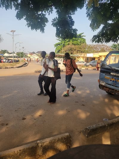The image depicts a street scene in San Martín, Colombia, under clear daylight conditions with visible shadows. Three individuals are walking from left to right across an asphalt road in the mid-ground. The leftmost subject is a male wearing a white t-shirt, dark trousers, and dark shoes, carrying a backpack. The middle subject is a female in a white and dark short-sleeved top, dark wide-leg trousers, and sandals, carrying a dark shoulder bag and holding a light-colored item. The rightmost subject is a female in an orange/brown patterned long-sleeved top, blue jeans, and light green sandals, also with a shoulder bag.
On the far right, a light blue Nissan van, displaying "NISSAN" and "MADU ABU CH" on its front, is partially visible, showing signs of rust on its bumper. In the immediate foreground, a concrete curb with white residue is present. The background features multiple solar-powered streetlights with rectangular panels, low-rise buildings, and extensive green foliage from trees. A gathering under a canopy with several other individuals is visible in the far-left background. Additional vehicles are present on the road in the distance. The overall environment suggests an urban or semi-urban area.