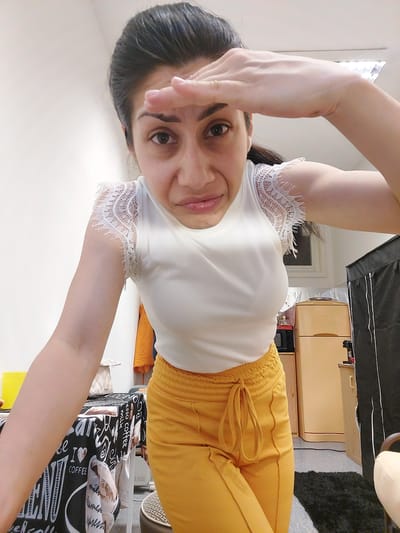 A medium shot captures a woman with dark hair pulled back, peering forward with her right hand shielding her eyes as if looking into the distance. She is wearing a white top with intricate white lace detailing on the cap sleeves and yellow drawstring pants. The background appears to be an indoor setting, possibly a room with a light-colored wall. A table covered with a black cloth featuring coffee-themed graphics is visible to the left, and a light-colored cabinet or appliance is seen to the right. There is no discernible connection to the city of Landshut, Germany.