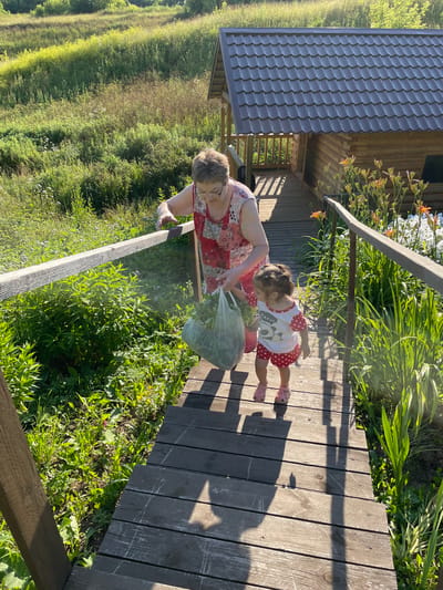 A woman and a young child are on a wooden walkway next to a building with a dark, wavy roof. The woman is wearing a red and white patterned dress, and the child is wearing a white shirt with red polka dots and red shoes. The child is holding a clear plastic bag filled with green leaves. They are surrounded by lush green vegetation and plants, some of which have orange flowers. The sun is shining, casting shadows on the wooden walkway, suggesting it is a sunny day. The scene appears to be outdoors, possibly in a garden or rural setting. The overall mood is peaceful and domestic. The image is oriented vertically.