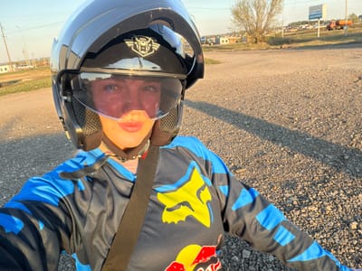 A person wearing a black, blue, and yellow Fox Racing shirt and a black helmet lies on a gravel surface. The shirt features a Red Bull logo and a Fox logo. A black strap crosses the person's body. The surrounding environment includes a grassy area, buildings, and a sign with Cyrillic text. The photo was taken in Азьмушкино, Russia.