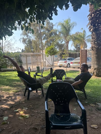The image portrays an outdoor scene in Nordia, Israel, under a clear blue sky, with the immediate foreground shaded by tree foliage. Two adult males are observed relaxing in dark plastic chairs on a patchy dirt and grass area.

The male on the left, dressed in a black t-shirt and olive green pants, is reclined with his right arm extended upwards and his left hand holding a smartphone, viewing its screen. His right leg is extended, resting on a second dark plastic chair. The male on the right, sporting a beard and wearing a black t-shirt and olive green pants, is also reclined in a dark plastic chair, looking towards the left. A small red and white object rests on the chair used as a footrest between them. An additional unoccupied dark plastic chair is positioned prominently in the foreground, with another further back on the left.

In the background, beyond a light-colored fence and gate, a paved area holds three parked vehicles: a white Seat Ibiza (license plate 70:610:01), a silver sedan (partially visible, license plate 70:868:01), and a white sedan. A green cylindrical waste bin is situated near the fence. The background landscape features numerous trees, including several tall palm trees, indicative of a semi-rural or suburban setting.