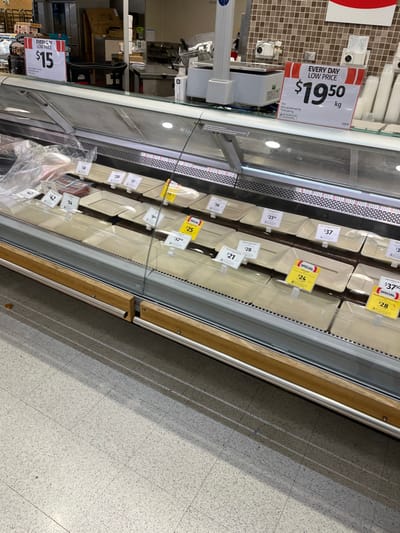 The image shows a refrigerated display case in a grocery store located in Middle Park, Australia. The case is filled with a variety of pre-packaged meat products. The products are labeled with prices, and the case is lit from above. There are no people present in the image.  The case is made of glass and stainless steel, and it is surrounded by a wooden frame. The case sits on a tiled floor. The case is also filled with items, including: a price sign, a meat thermometer, a metal rack, a plastic package with a chicken product, and various containers of meat products. The majority of the image is the refrigerated display case and the floor.