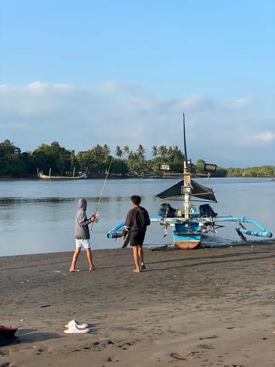 The image depicts a scene on the sandy bank of a river or estuary in Negara, Indonesia, under a clear, light blue sky with scattered white clouds, suggesting morning or late afternoon light. Two young males stand barefoot on the dark, damp sand.
The male on the left, wearing a grey hooded long-sleeve shirt and white shorts, holds a fishing rod with a reel, facing towards the water, appearing to be engaged in fishing. The male on the right, dressed in a dark long-sleeve shirt with white stripes on the sleeves and dark shorts, stands slightly behind and to the right of the first male, also facing the water and a nearby boat.
To their right, a traditional Indonesian outrigger fishing boat (Jukung) is partially beached on the dark sand, its stern facing the viewer. The boat's hull is blue on the upper section and reddish-brown below. It features blue outrigger poles extending laterally, equipped with blue and white striped cylindrical floats. Two outboard motors are mounted on the stern. A central mast rises from the boat, supporting a dark, partially furled sail and several rectangular floodlights. Two upper lights are mounted on a crossbar, while two others are positioned lower on the mast structure, with visible labels "200W 19VDC" on some.
The calm water of the river or estuary, appearing grey-blue, occupies the midground. Across the water, on the distant bank, a larger, ornate traditional wooden boat (Perahu) with high, curving ends is visible, either docked or partially ashore amidst dense tropical vegetation. Both banks of the waterway are lined with abundant trees, including numerous palm trees and other broadleaf species. On the immediate sandy bank, to the far left, a pair of white slippers or sandals are placed, with a portion of a red and black bag also visible. The sand exhibits various textures and footprints.