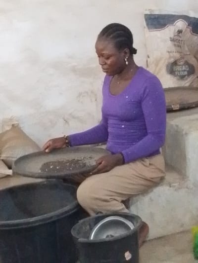 An adult female is depicted seated on a concrete ledge in an indoor setting in Olomoda, Nigeria. She has dark skin, braided hair, and wears a purple long-sleeved shirt and light brown trousers. She holds a dark, flat winnowing pan with both hands, containing a mixture of light granular material and dark specks, indicative of a sifting process. A large black plastic bucket is positioned below the pan to the left, and a smaller black bucket containing a metallic lid or bowl is to the right. In the background, a large sack marked 'DANGOTE BREAD FLOUR' rests against a rough, light-colored wall, alongside a smaller, lighter-colored sack.