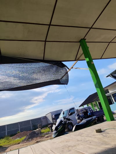 In the city of Dayahbaro, Indonesia, under a white paneled canopy supported by a bright green pillar, a black mesh shade is draped. The canopy's ceiling is composed of square panels with dark brown lines. In the background, a bright blue sky with scattered white clouds is visible.

Several vehicles are parked beneath the canopy. A white utility truck is parked next to a dark blue flatbed truck, which is loaded with what appears to be stacked mattresses. Two motorcycles are parked in front of the trucks. The motorcycle in the foreground is black and green, with its handlebars visible. The second motorcycle, positioned behind the first, is also black and blue.

To the left, a grey concrete wall stretches across the background, separating the paved area from a grassy field with piles of dirt and construction materials. Beyond the wall, a building with white walls and a dark roof is partially visible. A green pillar, similar to the one supporting the canopy, is attached to the side of this building.

On the paved ground beneath the canopy, a light brown electrical box is visible, connected to a green wire that runs across the concrete. The scene suggests an outdoor parking or storage area.