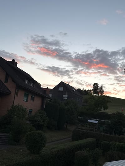 The image depicts a residential area in Lufingen, Switzerland, during a sunset. The sky is a soft gradient of pale blue and light grey, with streaks of pink and orange clouds. In the foreground, two houses are visible. The house on the left is a two-story brick building with a dark tiled roof and multiple windows. The house on the right is a darker, more modern structure with a gabled roof and several windows. Both houses are surrounded by lush greenery, including mature trees, bushes, and manicured hedges. A sloped, grassy hill is visible behind the houses, topped with several trees. The overall impression is of a peaceful, suburban evening in Lufingen.