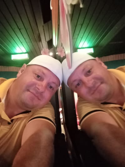 The image is a selfie of a man in a yellow collared shirt and a white baseball cap, taken indoors. The man's head and upper body are visible, and his reflection is also captured in what appears to be a mirrored surface. The background shows a wooden ceiling with green lighting. The man is smiling faintly and looking towards the camera. The location is not identifiable as being in the city of Hergiswil (NW), Switzerland.