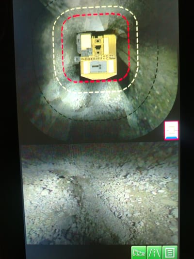 The image displays a split screen. The top portion shows a bird's-eye view of a construction vehicle, possibly a backhoe, within a circular path marked by dashed red and white lines on a gravelly surface. The vehicle is yellow and gray. A small, stylized icon is in the bottom right corner. The bottom portion of the screen shows a ground-level view of a rocky, uneven terrain, likely a construction site. In the bottom right corner of this lower section are three green icons, one resembling a bulldozer, another a road, and the third a document. The scene is not identifiable as being in Naminga, Russia.
