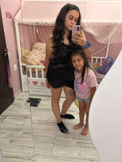 In a bedroom in Oreshak, Bulgaria, a young woman with dark, long hair takes a mirror selfie. She is wearing a black satin slip dress and black socks. Next to her stands a young girl, also with dark hair, wearing a pink t-shirt with a cartoon character and light-colored shorts. The girl is smiling at the camera. Behind them, a white bed frame with pink canopy netting is visible, adorned with small orange lights. A large teddy bear is seated on the bed. The room has light wood-paneled flooring and pink walls. A digital scale is placed on the floor in front of the mirror.