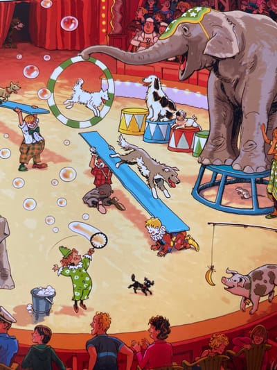 The image depicts a circus performance. In the background, a large gray elephant stands on a blue platform. In the mid-ground, a clown in a green polka-dot costume blows bubbles, and a dog jumps through a hoop held by another clown. Several other dogs and clowns are also present. In the foreground, an audience watches the show. The scene is set in a circus tent with red curtains and a yellow floor. There is no indication of the location being Edem, Russia.