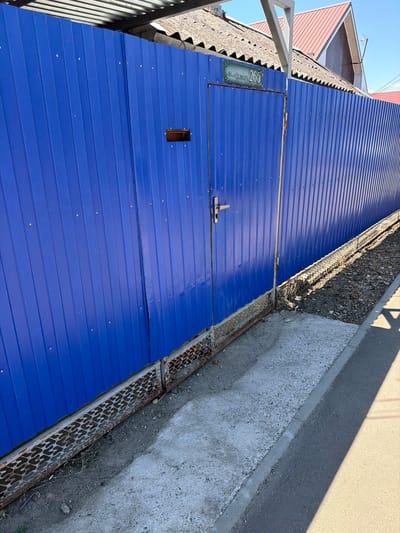 The image shows a blue corrugated metal fence with a gate. Above the gate, a sign reads "ул. 50 лет Октября 265". The fence is located on a street with a sidewalk and a grassy area. The sky is blue and the sun is shining, casting shadows on the ground. The houses visible in the background suggest a residential area. The overall scene depicts a street view in Timashevsk, Russia, likely in front of a property with the address indicated on the sign. There are no people or animals present. The condition of the fence appears to be generally good, though some rust is visible on the lower metal grate.