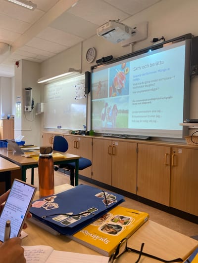 This image captures a classroom environment, likely in Uppsala, Sweden, given the Swedish text displayed on the projector screen. The room features typical educational elements: a large projector screen displaying text and images related to summer activities, a whiteboard with handwritten notes, a clock on the wall, fluorescent lighting, and student desks with learning materials. The projector is mounted on the ceiling. On one of the desks, a laptop is open, next to a water bottle, a blue folder adorned with stickers, and a yellow textbook. A smartphone is being held, displaying text and a keyboard, suggesting a student is engaged with their device. The desks and cabinets appear to be made of wood. The overall impression is that of an active classroom, possibly during a lesson or break.
