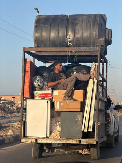 A heavily loaded white pickup truck with a custom-built, dark metal superstructure moves on an asphalt road in a-Zawayda, Palestinian Territory, under a clear blue sky.

Secured to the top of the metal frame is a large, black, horizontally oriented plastic water tank fitted with a green-handled faucet on its left end. Below this tank, within the metal frame, a young man with dark hair and a beard sits cross-legged on a pile of black bags and cardboard boxes. He wears a dark t-shirt and grey trousers, with brown sandals on his feet, and is holding a small dark object in his hands while looking to the right.

The cargo bed is densely packed with household items: a white refrigerator is positioned on the lower left. Above it, an orange-and-black boxed item, possibly a television, leans against the frame. A red and white box with Hebrew text sits atop another cardboard box. Multiple large black bags, various cardboard boxes, white bundled items, a dark grey metal cabinet, and several long white planks or profiles are also visible.

The background shows a dusty, semi-urban environment with rubble, partially constructed or damaged buildings, and utility poles with power lines under bright daylight.