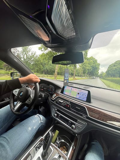 An interior shot of a modern BMW car shows the driver's perspective from the front passenger seat, looking towards the road ahead. The image is oriented sideways, with the driver's left hand on the steering wheel and their legs, dressed in ripped blue jeans, visible in the lower right.
The car's dashboard features a prominent infotainment screen, and to its right, a smartphone is mounted vertically, displaying navigation information. The phone's screen shows a map, current speed (52 km/h) against a 50 km/h limit, an instruction for "800 m" to "Wijkertaan," and a destination named "Jolanda voorburg" 1.2 km away with an estimated arrival between "10:00-11:00." Media controls are also visible on the phone.
The car's interior features wood trim on the dashboard and center console. A climate control panel shows a temperature of "22.0" degrees Celsius with "AUTO" mode active. Coins are visible in a cup holder, next to a green cylindrical object. An air freshener with the number "22" hangs from the rearview mirror.
Through the windshield, a road lined with trees is visible under an overcast sky, suggesting daytime driving. Other vehicles can be seen in the distance. The text "AIRBAG" is visible on the passenger side pillar. The main car display also shows vehicle speed (54 km/h) and other driving data. The overall scene depicts a person driving on a road, likely using the navigation system for directions to a location in Voorburg, Netherlands.