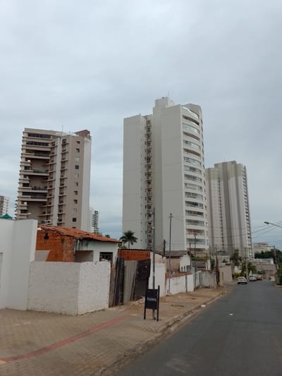 The image depicts a street scene in Cuiabá, Brazil, under an overcast sky. In the foreground, a paved sidewalk borders an asphalt road. A white, textured wall with a red stripe runs along the sidewalk. A small, dark sign with white text, possibly a street sign or directional marker, stands on the sidewalk. Behind the wall, a low-rise structure with a red-tiled roof and brick walls is visible, appearing to be under construction or in disrepair.

Dominating the background are several tall, modern residential buildings. The tallest building, on the left, is a light-colored high-rise with numerous balconies and staggered architectural elements. To its right are two more slender, light-colored high-rise apartment buildings, one with curved balconies. Further in the distance, other buildings and trees are discernible, hinting at a developed urban environment. A few cars are parked along the street, and power lines are strung between utility poles. The overall impression is of a residential neighborhood in a Brazilian city.
