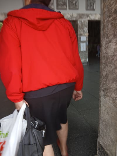 A person is captured from behind, wearing a red hooded jacket over a black outfit, and is holding a white plastic bag and a black leather handbag. The person appears to be walking towards an entrance, possibly a building, with a textured stone wall to the right and a darker interior visible through an archway. The ground is paved with dark tiles. The image suggests an urban setting, potentially a public space in Truskavets, Ukraine, given the presence of a person with shopping bags and the architectural style of the surrounding environment. The overall scene is slightly blurred, indicating movement or a lower-resolution capture.