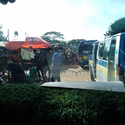 The image depicts an active open-air market and transport hub in Mukuyuni, Kenya, captured from the interior of a vehicle. In the foreground, a dark green, textured surface, possibly artificial grass on a dashboard, dominates the lower portion, with a light-colored, flat object visible directly above it.
The midground features an informal marketplace on the left, characterized by makeshift stalls and an orange sun umbrella with "SAFARICOM" branding. Underneath the umbrella, individuals are present, with one person in dark attire seated and another standing near green bundles on the ground. Several people are walking through the central area, including one man prominently visible in a blue shirt and a light-colored cap, facing away from the viewer. The ground throughout this area appears unpaved, with some metal grate-like structures visible on the dirt.
To the right, two passenger vans, commonly known as matatus, are present. The closest van on the right is blue and silver/white, with its side door open and luggage secured on its roof rack. Behind it, a dark blue van marked "PRESTIGE" is visible, displaying a Kenyan license plate (e.g., "K** 205"). People are congregated around and between these vehicles, suggesting passenger movement or commercial activity.
In the background, a water tower structure is visible on the left horizon, with numerous trees and foliage indicating a mix of natural and built environments. The sky is bright with partial cloud cover.