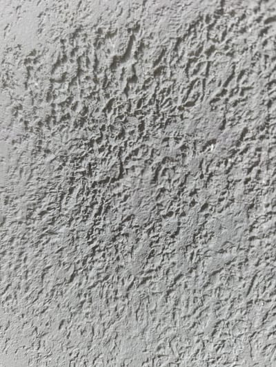 The image is a close-up, full frame, vertically oriented shot of a textured, light gray surface, possibly a wall or ceiling. The texture is rough and uneven, with a pattern of small, irregular bumps and indentations that create shadows and highlights. The lighting appears to be coming from the top left, casting shadows on the lower right portions of the textured areas. There are no people, objects, or identifiable landmarks present in the image. The background is entirely this textured surface. The image does not depict any specific location within the city of Uchquduq, Uzbekistan, or any urban environment. It is a purely abstract textural study.