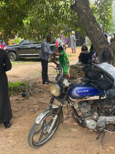 A medium shot shows an outdoor scene in New Nyanya, Nigeria. In the foreground, a blue and white motorcycle is parked. The motorcycle has a single headlight, a chrome fender, and a black seat. On the gas tank, the word "QLINK" is visible. A black bag is strapped to the back of the motorcycle.
To the left of the motorcycle, a person in a long black robe is standing, facing away from the camera. To the right of the motorcycle, a person wearing a white t-shirt is seated, facing away from the camera.
In the middle ground, a man in a grey jacket and black pants is shaking hands with a woman wearing a green and black patterned dress. Behind them, a black SUV is parked. Several other people are visible in the background, some standing and some seated, under the shade of a large tree. The ground is covered in dirt and fallen leaves. The general atmosphere appears to be a gathering or event.