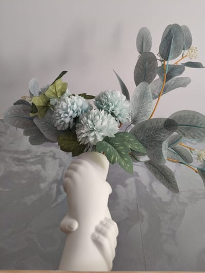 A white ceramic vase shaped like clasped hands holds a bouquet of artificial flowers and eucalyptus branches against a light gray background. The flowers are a pale blue, clustered and pom-pom shaped. The eucalyptus leaves are a dusty blue-green with visible vein patterns. The vase is positioned on a wooden surface. The background features a wall with a subtle textured pattern. There are no people or animals present in the image. The scene depicts a still life arrangement. The location in Shimanovsk, Russia is not discernible from the image.