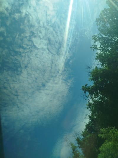 The image captures a low-angle view of a bright, cloudy sky with sun rays piercing through. The sky is a vibrant blue with scattered white clouds that appear textured. On the right side of the frame, the tops of lush green trees are visible, with some bare branches interspersed among the foliage. The sunlight creates a strong lens flare effect, particularly in the upper portion of the image, casting bright streaks of light. The perspective suggests the photograph was taken from the ground, looking upwards, likely within a natural or semi-urban environment like the city of Rusca, Moldova, where trees are prevalent and open sky is visible. There are no people or specific landmarks visible to pinpoint an exact location within Rusca.