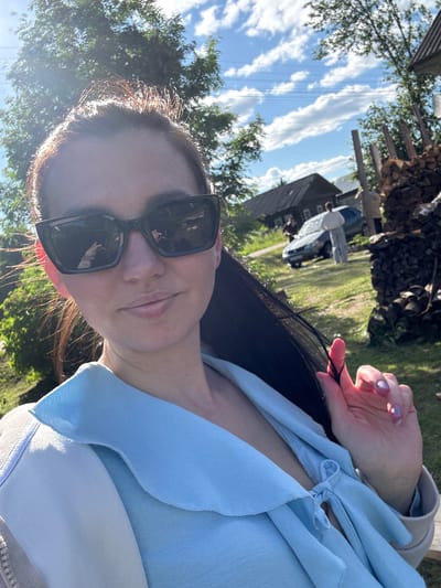 The image is a selfie taken by a young woman outdoors, rotated to a vertical orientation in the file but depicting a horizontal scene. The woman is the primary subject, positioned in the foreground and looking directly at the camera with a relaxed expression. She has long, dark hair, visibly illuminated by strong sunlight, and is wearing dark, rectangular sunglasses. Her attire includes a light blue top with a ruffled or layered neckline, possibly tied at the chest, worn over a white undershirt. Her left hand, adorned with light, shimmery nail polish, is gently touching a strand of her hair.
The background suggests a rural or semi-rural setting in Луптюг, Russia. The sky is bright blue with scattered white clouds, indicating clear and sunny weather during daytime. Green foliage from trees frames the scene and extends into the mid-ground. Visible in the distance are several structures: a dark wooden house or building with multiple windows, a light-colored car (possibly white or light blue) parked near the building, and a large woodpile stacked beneath an overhang or shed. Green grass covers the ground in the background. Thin, dark vertical lines, possibly power lines, are faintly visible against the sky. The strong sunlight creates a luminous effect on the woman's hair and clothing. No text is visible in the image.