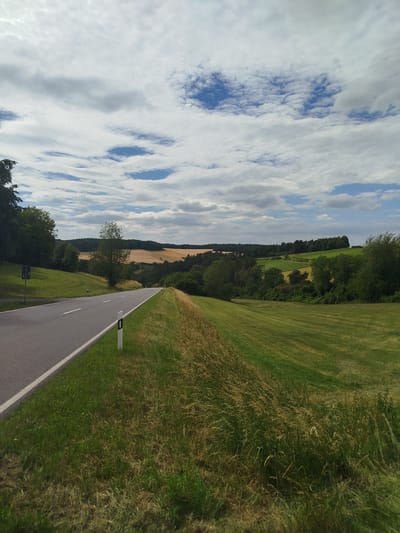 This media file captures a scenic view of a rural road in Bad Mergentheim, Germany. The road, paved and with a white lane marking, curves gently into the distance, bordered by lush green grassy fields. The fields are interspersed with trees and further back, rolling hills are covered in a patchwork of green and golden agricultural fields, suggesting a landscape of farms and meadows.

The sky is a dominant feature, filled with a dramatic display of clouds. Large, fluffy white clouds drift across a bright blue sky, with some areas appearing more grey and overcast, indicating a dynamic weather pattern. Sunlight filters through the clouds, casting soft light on the landscape.

A white road marker with black bands is visible along the side of the road, as is a dark rectangular sign on a post to the left. The overall impression is one of a serene and picturesque countryside setting, likely during daytime, given the brightness of the scene. There are no visible people or animals in the frame. The image evokes a sense of peace and natural beauty.