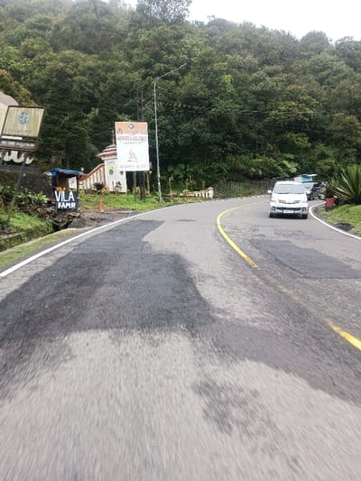 The image displays a winding, wet asphalt road through a densely forested, mountainous region. Signs visible in the image, specifically "VILA KAMR" and "BUMI AKI," indicate Indonesian language, contradicting the specified location of Deothang, Bhutan.
The road features a solid yellow center line for the right-hand lane and a solid white line marking the left edge, with patches of moisture across its surface. On the left side of the road, a small, white, decorative building with a red-tiled roof and balustrades stands, possibly an entrance or gatehouse. Adjacent to it, a large vertical sign displays text including "MENORAH & JAY GANTIYA" and "BUMI AKI," with a smaller sign below it showing a local emblem. Closer to the foreground on the left, another sign reads "VILA KAMR." A utility pole with a streetlight is positioned near these signs. The immediate left environment consists of dark soil and dense, green vegetation, including small cultivated plants.
On the right side of the road, a silver minivan is visible driving away from the camera, having just rounded a gentle right curve. Behind it and further up the road, a blue vehicle (possibly a utility truck with a canopy) is partially visible. The right side of the road is bordered by lush green foliage and a sloped terrain. The background consists of steep, thickly forested hills under an overcast sky. No individuals are visible in the scene.