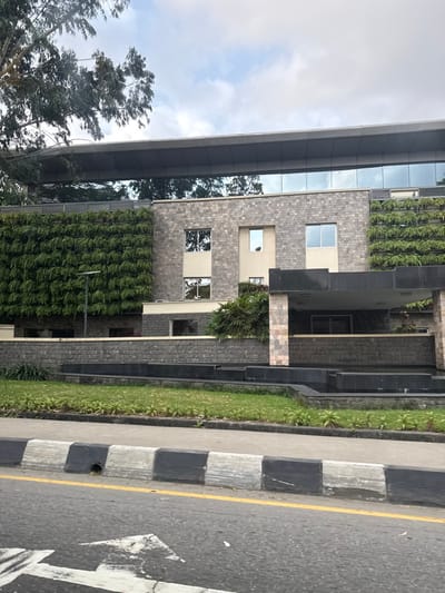 The image displays a modern multi-story building located in Falomo, Nigeria, observed from a street-level perspective. The building's facade is constructed from light-colored, rectangular stone blocks and features two prominent vertical green walls densely covered with foliage. An upper section of the building incorporates a reflective glass curtain wall beneath a flat, overhanging gray roof structure. Several rectangular windows are visible, reflecting the cloudy sky and nearby trees.

At the ground level, a dark-colored canopy extends from the building, supported by stone elements. The immediate landscaping includes tiered dark stone retaining walls, what appears to be a dry water feature or decorative planter, and manicured green lawns planted with small, uniform flora.

In the foreground, a gray asphalt road is visible, marked with a continuous yellow line near the curb and a faded white arrow indicating direction. The concrete curb is painted with alternating black and white stripes. A large tree with green leaves partially frames the upper left side of the scene. No individuals or discernible activities are present. The sky is overcast with visible cloud formations.
