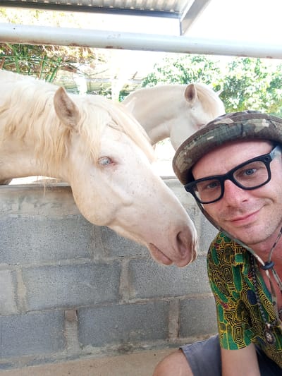 A man wearing a camouflage hat, glasses, and a patterned shirt is taking a selfie with two horses at a stable. The man is smiling and looking at the camera. The horses are white and appear to be at a stable. The background shows trees and a metal roof. The location is Phước Hải, Vietnam.