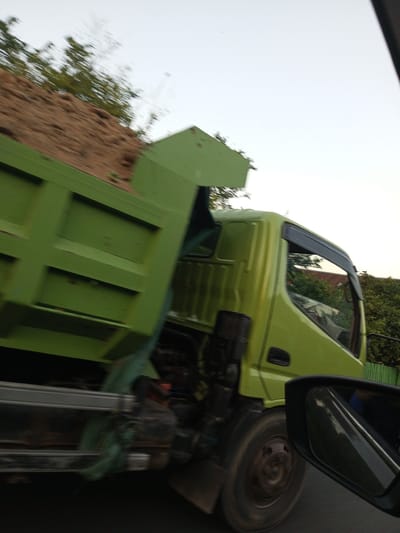 In Pallawarukka, Indonesia, a light green dump truck is captured in motion, its cargo bed piled high with brown dirt. The truck's cab is also light green, featuring a dark tinted window with a visible rain guard. A black side mirror reflects a partial view of the truck and the surrounding environment. The truck's wheel is visible, appearing worn. In the background, lush green trees and a reddish-brown building or structure can be seen. The sky is a pale, overcast white. The image has a motion blur, suggesting the vehicles were traveling at a significant speed.